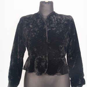 Op de afbeelding: Een zwart fluwelen jasje met lange mouwen, een gestructureerde kraagloze halslijn en een peplum-zoom. De stof heeft een gekreukte textuur over het hele oppervlak en het kledingstuk sluit aan de voorzijde met een enkele knoop in de taille.