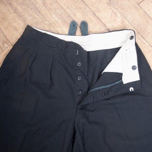 Puede incluir: Pantalones negros con bragueta de botones y cinturilla blanca. Los pantalones tienen pliegues en la parte delantera y dos pequeñas trabillas. La bragueta de botones está parcialmente desabrochada, revelando el interior. La tela parece ser un material oscuro y resistente.