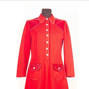 Pode incluir: Um vestido vermelho brilhante de manga comprida com gola pontiaguda, uma carcela de botões vertical com botões brancos e dois bolsos frontais com abas. A peça inclui costuras decorativas brancas ao longo dos ombros e das bordas dos bolsos.
