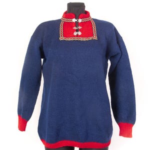 Könnte beinhalten: Ein dunkelblauer Langarmpullover mit rotem Kragen, roten Bündchen und rotem Saum. Der Brustbereich zeigt ein rotes Paneel mit einer dekorativen gelb-grünen geometrischen Bordüre, das am Halsausschnitt mit zwei silberfarbenen Metallschließen versehen ist.