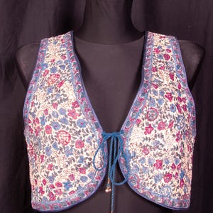 Puede incluir: Un chaleco sin mangas con una base de color crema y un denso estampado floral en tonos rosa, azul y gris. La prenda presenta un escote en pico profundo, ribetes azules en los bordes y un cierre frontal de lazo con dos pequeños dijes metálicos.