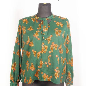 Op de afbeelding: Smaragdgroene blouse met lange mouwen en een bloemenpatroon in oranje en bruin. De blouse heeft een sleutelgat-halslijn en een stropdas. De stof lijkt lichtgewicht, geschikt voor warmer weer.