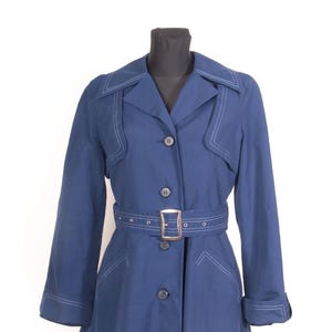 May include: A navy blue trench coat with white stitching details. The coat features a belted waist with a silver buckle, a notched collar, and button closures. The sleeves are long, and the coat is displayed on a black mannequin.