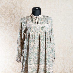 Puede incluir: Vestido de manga larga con estampado floral en tonos verdes, beige y amarillo. El vestido presenta un cuello alto con detalle de lazo y ribete de encaje. El vestido se exhibe en un maniquí negro.