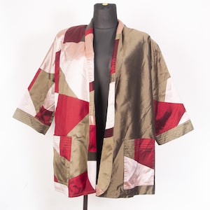 Könnte beinhalten: Kimono-Jacke im Patchwork-Stil in Rot-, Rosa- und Olivgrüntönen. Die Jacke hat ein geometrisches Muster mit zwei Vordertaschen und weiten Ärmeln. Das Kleidungsstück wird auf einer schwarzen Schaufensterpuppe präsentiert.