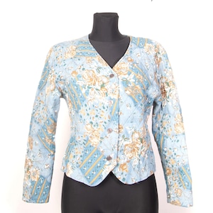 Op de afbeelding: Lichtblauwe gewatteerde jas met een bloemenpatroon in geel, wit en groen. De jas heeft een V-hals, lange mouwen en drie bruine knopen. De jas wordt gepresenteerd op een zwarte mannequin. Vintage stijl.