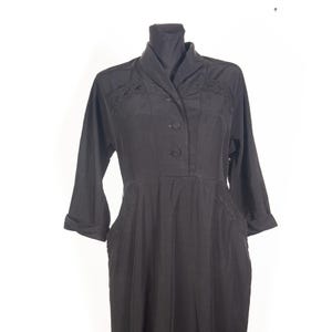 May include: A black long-sleeved dress featuring a shawl collar, a three-button front closure, and decorative tonal embroidery detailing on the shoulders and waistline. The garment has cuffed sleeves and a structured bodice design.
