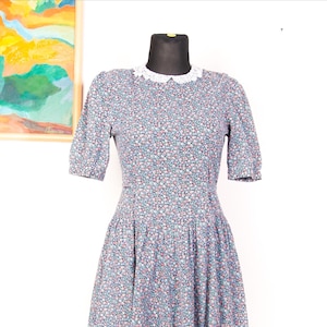 Op de afbeelding: Een knielange jurk met een donkerblauwe basis en een dicht bloemenpatroon van kleine roze, witte en blauwe bloemetjes. Het kledingstuk heeft korte mouwen, een aansluitend lijfje, een geplooide taille en een witte kanten kraag met schulprand.