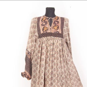 Puede incluir: Vestido beige de manga larga con estampado floral y escote de ojo de cerradura. El vestido presenta intrincados bordados en tonos marrones y dorados alrededor del escote y en las mangas. El vestido se exhibe en un maniquí negro.