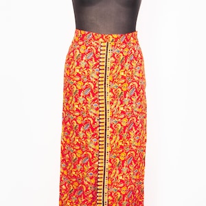 May include: A long, red skirt with a vibrant floral pattern in yellow, blue, and green. The skirt features a central black stripe with yellow and gold detailing. The skirt is displayed on a mannequin.