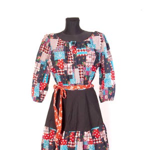 Può includere: Un abito patchwork con maniche lunghe e gonna nera. La parte superiore presenta un mix di toppe in tessuto floreale, a quadri e tinta unita in rosso, blu e rosa. Una cintura a pois rossi e bianchi stringe la vita.