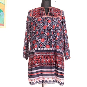 Puede incluir: Una túnica de manga larga con un estampado floral en rojo, blanco y gris sobre un fondo azul marino. El vestido tiene un escote decorativo y un borde estampado en el dobladillo. El vestido se exhibe en un maniquí.
