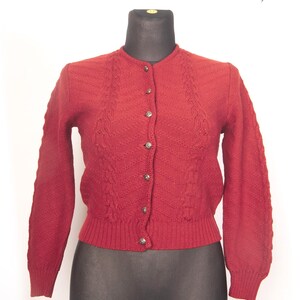 Puede incluir: Un cárdigan rojo de estilo vintage, con cuello redondo y mangas largas. El suéter presenta un patrón de punto texturizado y se abrocha con cinco botones plateados en la parte delantera. La prenda se exhibe en un maniquí negro.