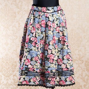 May include: A knee-length skirt featuring a black background with a dense floral pattern in shades of pink, light blue, and cream. The skirt has vertical pleats, a fitted waistband, and three horizontal black bands above a scalloped black lace hem.