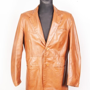 以下が含まれることがあります： 2つボタン留め、ノッチドラペル、フロントポケット2つのタンレザージャケット。黒いマネキンに展示されており、クラシックでテーラードなデザインです。ブレザーはミディアムブラウン色です。