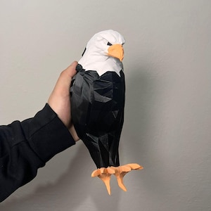 Puede incluir: Una escultura geométrica low-poly de un águila calva con cabeza blanca, cuerpo negro y pico y garras de color naranja. La figura está construida con facetas angulares afiladas que crean una apariencia facetada distintiva en toda el ave.