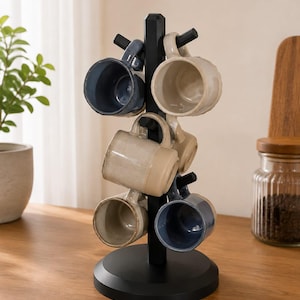 Puede incluir: Un soporte para tazas de metal negro con base circular y seis ganchos en ángulo, que sostiene una colección de tazas de cerámica en tonos azul, crema y beige. Las tazas tienen un acabado esmaltado liso y están dispuestas alrededor del poste central.