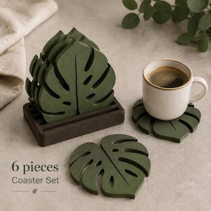 Puede incluir: Un juego de seis posavasos de color verde oscuro con forma de hojas de monstera, apilados en un soporte rectangular marrón oscuro, con un posavasos plano y una taza de cerámica blanca con café. El texto dice 6 pieces Coaster Set.
