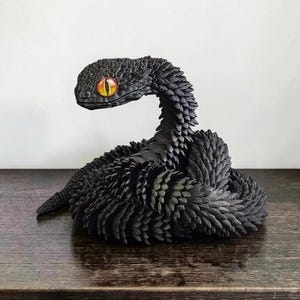 Könnte beinhalten: Eine schwarze Schlangenfigur mit einer stark texturierten, schuppenartigen Oberfläche, die den gesamten gewundenen Körper bedeckt. Die Schlange hat ein markantes, realistisches Auge mit leuchtend orangefarbener Iris und vertikaler Pupille.