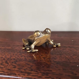 Puede incluir: Una pequeña figura de rana hecha de latón pulido. La rana está representada en una posición sentada y de descanso, con las patas delanteras extendidas hacia adelante y sus grandes ojos redondos mirando hacia arriba, mostrando un tono dorado.