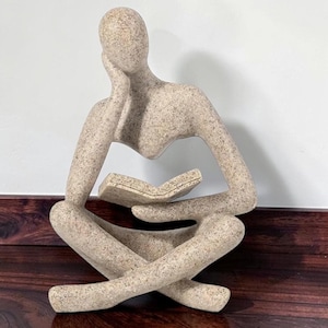 Puede incluir: Una escultura de piedra beige moteada que representa una figura sentada con las piernas cruzadas. La figura tiene una mano apoyada contra la cabeza y la otra mano sosteniendo un libro abierto. La superficie tiene un aspecto texturizado y granulado.
