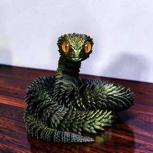 May include: A coiled snake figurine featuring a metallic dark green, scale-like textured body and large, prominent orange eyes. The articulated sculpture has a detailed, layered appearance with a pointed head and a curved, segmented tail structure.