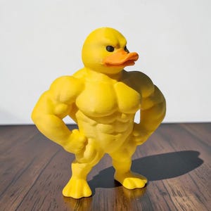 Pode incluir: Uma estatueta de pato amarelo com um físico musculoso semelhante ao humano, apresentando músculos peitorais, abdominais e braços definidos. O pato tem um bico laranja brilhante e pequenos olhos pretos, numa postura ereta e confiante.