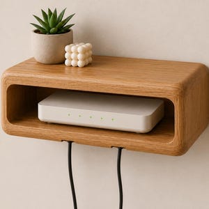 Puede incluir: Un estante de pared rectangular de madera marrón claro con esquinas redondeadas y un compartimento central abierto. Un router electrónico blanco se encuentra dentro del estante, mientras que una pequeña suculenta verde y una vela blanca texturizada están encima.