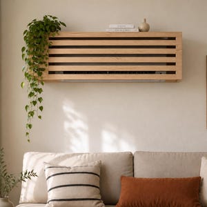 May include: A light-colored wooden slatted cover mounted on a wall over an air conditioning unit. The rectangular cover features horizontal slats and a flat top surface holding two books and a small vase. A trailing green plant hangs from the left side.
