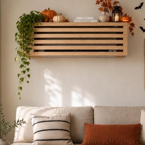 May include: A light-colored wooden slat cover for a wall-mounted air conditioning unit. The rectangular frame features horizontal wooden bars and is mounted on a wall above a sofa, serving as a shelf for decorative items like pumpkins and a lantern.