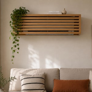May include: A rectangular light brown wooden slatted cover mounted on a wall, designed to conceal an air conditioning unit. The top surface of the cover functions as a shelf, holding two white books and a small beige ceramic vase.