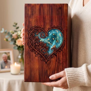 Könnte beinhalten: Eine rechteckige Urne aus Holz mit einer dunkelbraunen Holzmaserung. Die Vorderseite zeigt eine große, kunstvolle herzförmige Einlage, die mit wirbelndem türkisfarbenem und blauem Harz gefüllt und mit kleinen Blattgoldpartikeln verziert ist.