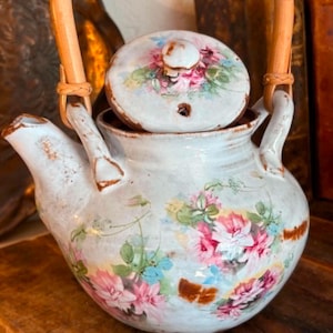 Könnte beinhalten: Eine Keramik-Teekanne mit hellblauer Glasur und einem Blumenmuster aus rosa und weißen Rosen. Die Teekanne hat einen geschwungenen Ausguss, einen passenden Deckel mit einem kleinen Knauf und einen abgerundeten Henkel aus hellem Rattangeflecht.