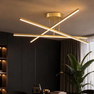 Peut inclure: Un plafonnier doré composé de trois barres LED linéaires entrecroisées diffusant une lumière chaude. Le luminaire est fixé au plafond par une base circulaire et de fines tiges verticales, créant un motif lumineux géométrique multidirectionnel.