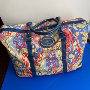 May include: A large travel bag featuring a vibrant paisley pattern in shades of red, yellow, blue, and cream. The bag includes dark blue leather handles, a top zipper closure with heart-shaped pulls, and a central oval logo patch on the front.