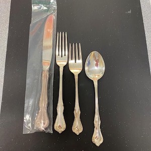 May include: A set of silver-toned flatware consisting of a dinner knife sealed in a clear plastic sleeve, two forks, and a spoon. Each piece features a decorative, ornate handle with a polished metallic finish and intricate detailing at the base.