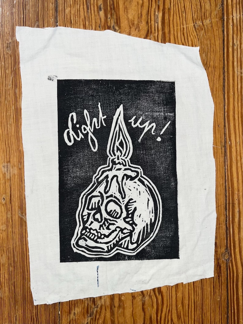 Pu&ograve; includere: Un pezzo rettangolare di tessuto bianco con un'illustrazione stampata in nero di un teschio umano con una candela accesa che si scioglie sopra. Il testo "light up!" &egrave; stampato in corsivo bianco sopra la candela entro il bordo nero.