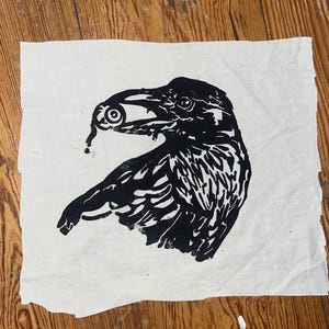Könnte beinhalten: Ein quadratisches Stück cremefarbenen Stoffes mit einem schwarzen Linoldruck eines Raben im Profil. Der Vogel hält einen menschlichen Augapfel im Schnabel, wobei ein kleiner Tropfen Flüssigkeit vom Auge nach unten abfällt.