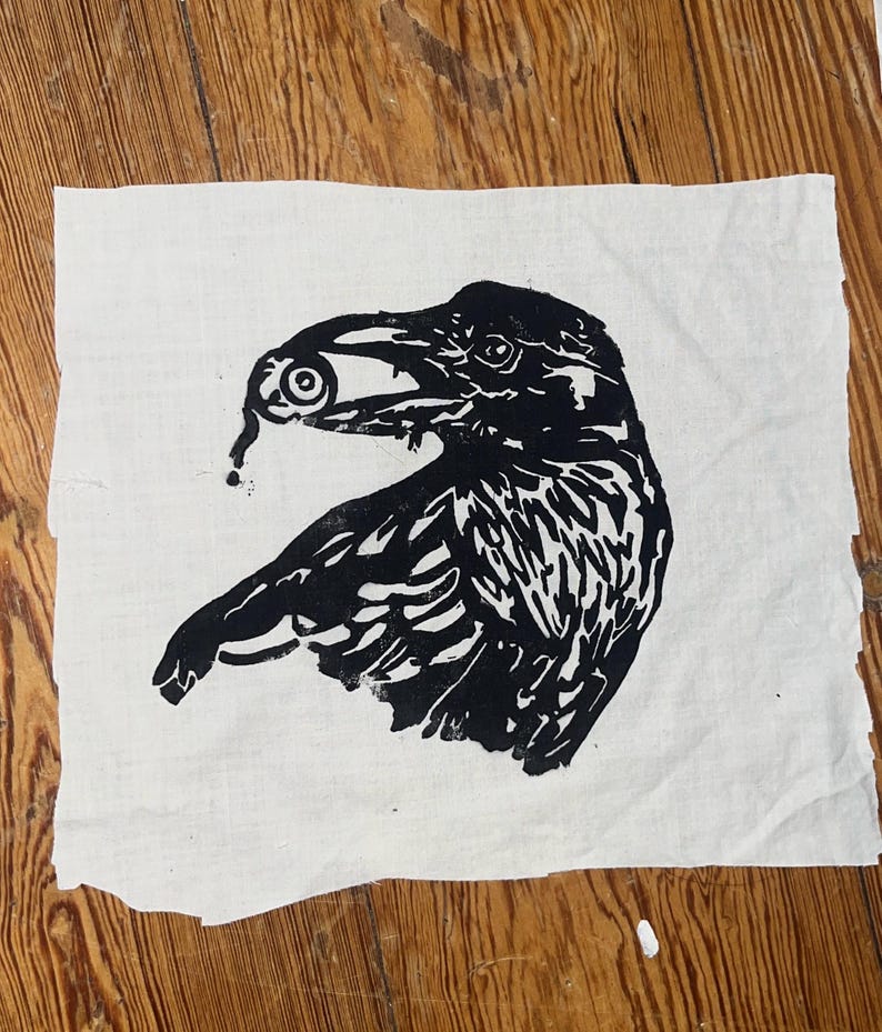 May include: A square piece of off-white fabric featuring a black ink block print of a raven in profile. The bird is depicted holding a single human eyeball in its beak, with a small drop of liquid trailing downward from the eye.
