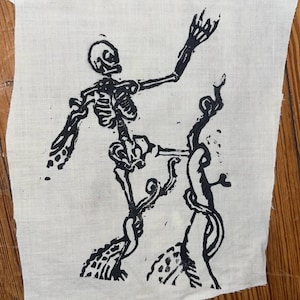 Könnte beinhalten: Ein rechteckiges Stück cremefarbenen Stoffes mit einem schwarzen Blockdruck eines Skeletts, das auf einem Bein steht. Das Skelett ist mit einem erhobenen Arm und einem angewinkelten Bein dargestellt und interagiert mit einer hohen, gewundenen Struktur.