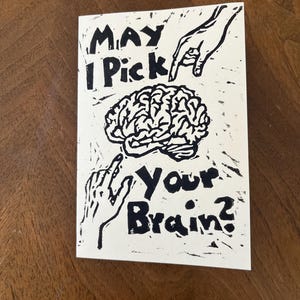 Könnte beinhalten: Eine rechteckige Grußkarte mit einem schwarzen Blockdruck auf weißem Hintergrund. Das Motiv zeigt ein menschliches Gehirn in der Mitte, auf das zwei Hände zeigen. Der Text lautet, May I Pick Your Brain? in einem kräftigen, handgezeichneten Stil.