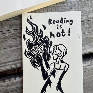 Könnte beinhalten: Eine rechteckige weiße Karte mit einer schwarzen Tuschezeichnung einer Person, die ein brennendes Buch hält. Der Text Reading is hot! ist in schwarzer Tinte über der Figur gedruckt. Das Design ist in einem kräftigen Linolschnitt-Stil gehalten.