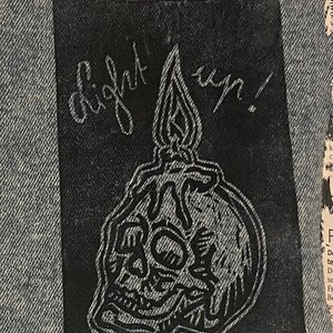 Pu&ograve; includere: Una toppa rettangolare in tessuto nero con un'illustrazione a linee bianche di un teschio umano con sopra una candela che si scioglie. Il testo "light up!" &egrave; scritto in corsivo sopra la candela. La toppa &egrave; cucita su un tessuto denim azzurro.