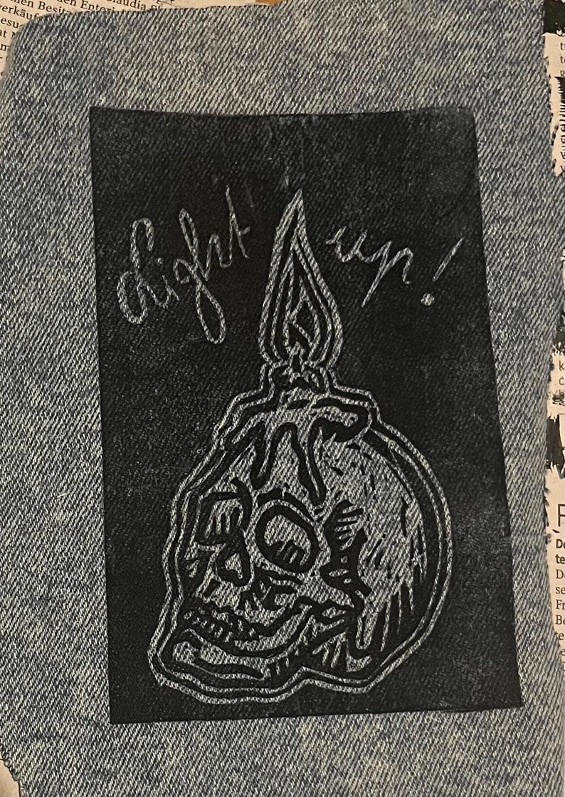Pu&ograve; includere: Una toppa rettangolare in tessuto nero con un'illustrazione a linee bianche di un teschio umano con sopra una candela che si scioglie. Il testo "light up!" &egrave; scritto in corsivo sopra la candela. La toppa &egrave; cucita su un tessuto denim azzurro.