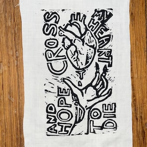 Könnte beinhalten: Ein rechteckiges weißes Stoffstück mit einem schwarzen Blockdruck-Design, das ein anatomisches Herz und eine Hand zeigt. Der Text CROSS MY HEART AND HOPE TO DIE ist um die zentralen Illustrationen mit einer strukturierten Optik angeordnet.