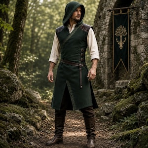May include: A dark green hooded tunic featuring a wrap-style front with intricate embroidery, a brown leather belt, and side buckle closures. The garment is worn over a cream-colored long-sleeve shirt, paired with dark trousers and brown leather boots.