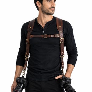 May include: A dark brown leather dual camera harness featuring adjustable straps, brass-toned metal buckles, and swivel clips. The harness is worn over a black long-sleeve henley shirt and black trousers, supporting two professional cameras at the hips.