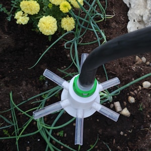 Pode incluir: Uma cabeça de aspersor de jardim de plástico branco com seis bicos tubulares transparentes e um anel de bloqueio verde, ligada a uma mangueira de borracha preta. O texto "fatto a strati" é visível no canto superior esquerdo.