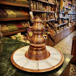 Peut inclure: Une théière en cuivre à deux étages avec des gravures florales complexes et des poignées en bois foncé, reposant sur une base circulaire en cuivre entourée de dix petits plats en céramique blanche trapézoïdaux disposés en cercle.
