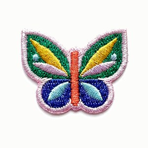 May include: An embroidered butterfly patch featuring a central orange body. The upper wings are green with yellow and light purple accents, while the lower wings are dark blue with light blue details. The entire patch is outlined in light pink thread.